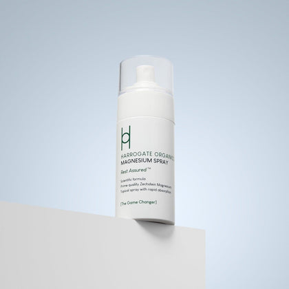 A white bottle of Harrogate Organics Magnesium Spray - 100ml sits on a white surface with a pale blue background. The label reads "Rest Assured" and highlights organic wellness and rapid absorption benefits.