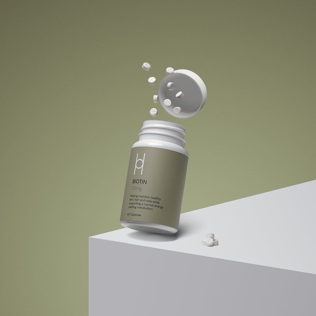 A bottle of biotin supplements with its cap open, white tablets floating in the air, and a few tablets resting on a white surface against a muted green background.