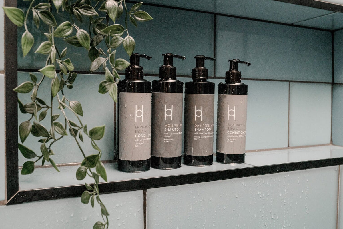 Four black bottles of hair care products with pumps are lined up on a shower shelf, next to a hanging leafy green plant, against a blue tiled wall with water droplets.
