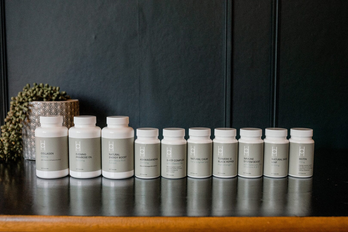 Ten white supplement bottles with gray labels are lined up on a black surface in front of a dark wall. A small plant in a patterned pot is partially visible on the left.