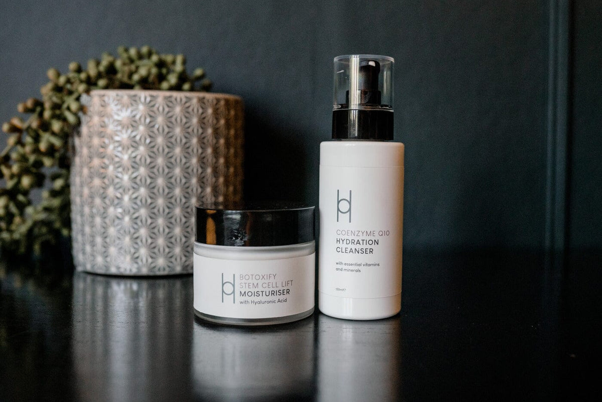 Two white skincare products on a dark surface: a jar labeled Botoxify Stem Cell Lift Moisturiser and a pump bottle labeled Coenzyme Q10 Hydration Cleanser, with a textured candle and greenery in the background.