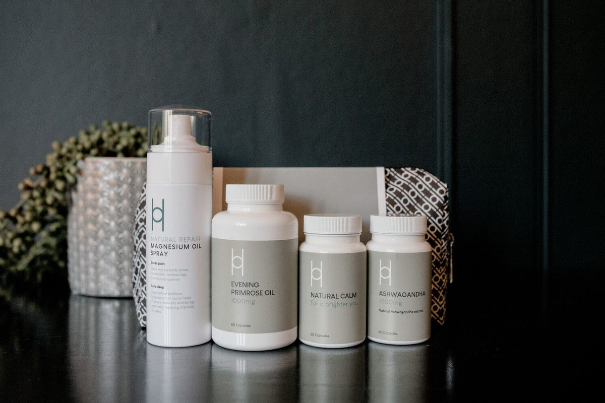 Four white bottles of wellness products, including magnesium oil spray, evening primrose oil, natural calm, and ashwagandha, are arranged on a dark surface with a decorative box and plant in the background.