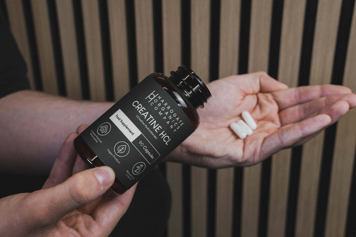 A person holds a bottle of Hydrolyze Performance Lab Creatine HCL capsules in one hand and two white capsules in the other, against a backdrop of vertical wooden slats.