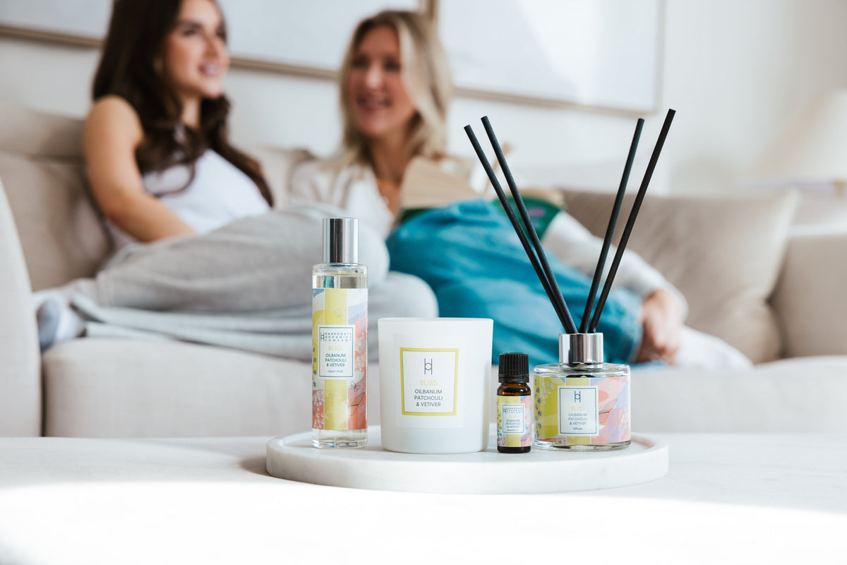 Two women sit on a couch in the background, smiling and talking, while a tray with scented products like a candle, diffuser, spray, and oil is displayed in focus in the foreground.