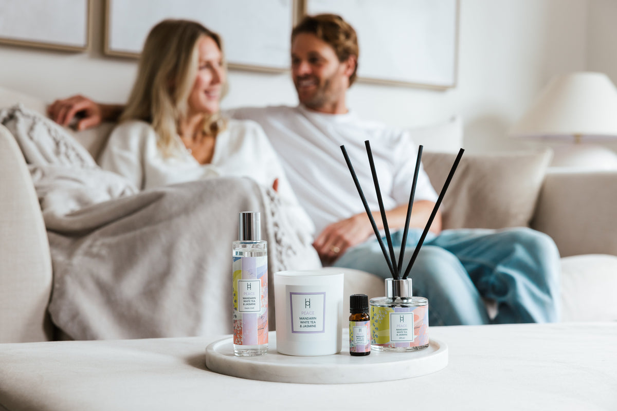Two women sit on a couch in the background, smiling and talking, while a tray with scented products like a candle, diffuser, spray, and oil is displayed in focus in the foreground.