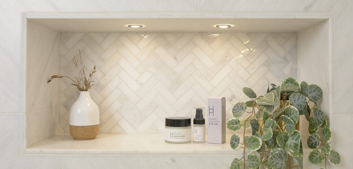 A recessed bathroom shelf with herringbone tile contains a white vase with dried plants, a potted green plant, a jar, a pump bottle, and a skincare product box under two small spotlights.