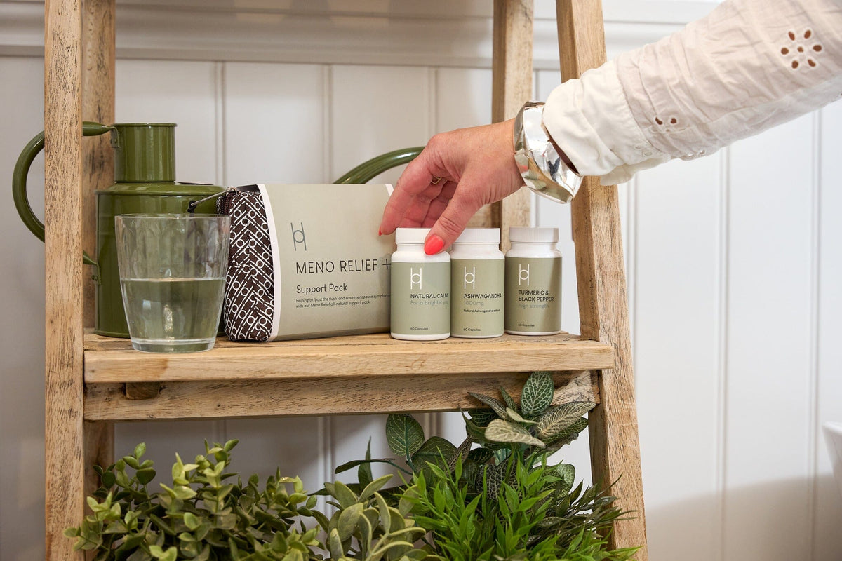 A hand reaches for a supplement bottle on a wooden shelf holding a glass of water, a green teapot, a Meno Relief Support Pack, and various potted plants.