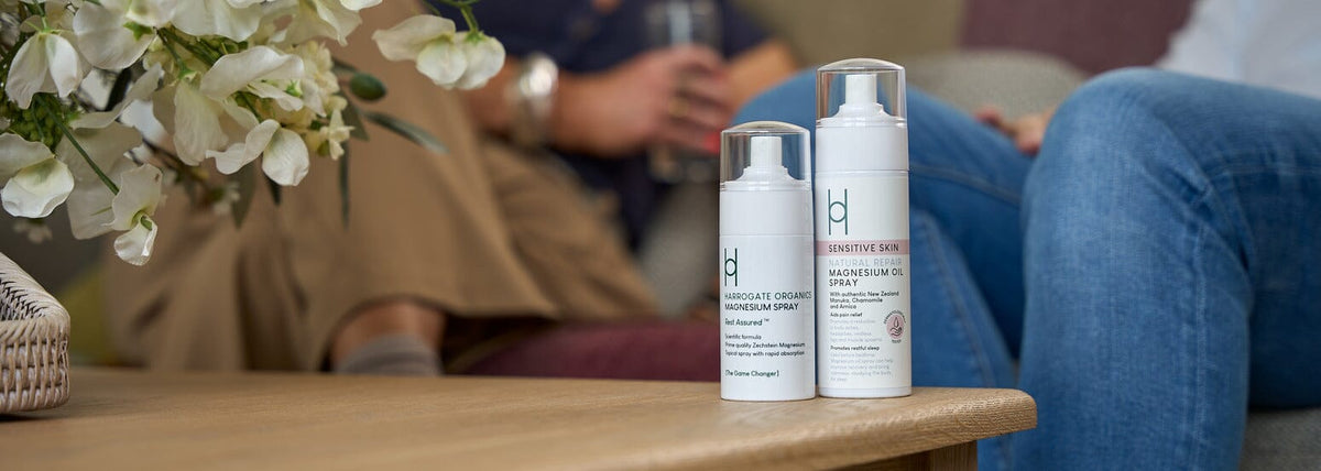 Two bottles of skincare spray labeled “Sensitive Skin” sit on a wooden table, with blurred people sitting in the background and white flowers visible to the left.