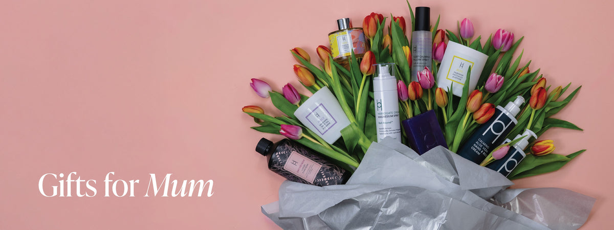 Mother’s Day Gifting text on a pink background with colorful tulip bouquets and assorted gift items like candles, chocolate, wine, and perfume arranged on both sides.