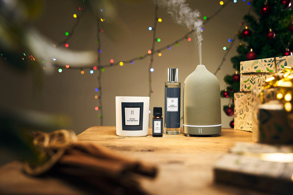 A candle, essential oil bottle, spray, and diffuser sit on a wooden table with wrapped gifts, a Christmas tree, and colorful string lights in the background.