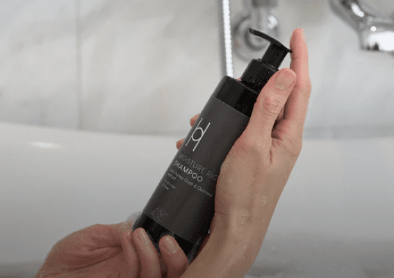 A person holds a black bottle of shampoo with a pump dispenser over a white bathtub. The label reads Moisture Rich Shampoo.
