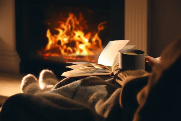 A person wrapped in a blanket sits by a cozy fireplace, holding a mug and an open book, with flames burning warmly in the background.