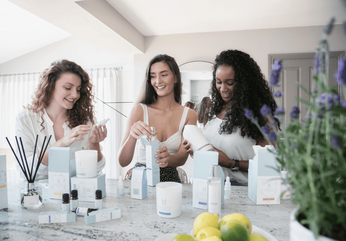 Three women smiling and using skincare products together at a kitchen counter, surrounded by candles, beauty items, and fresh lemons, with purple flowers in the foreground.