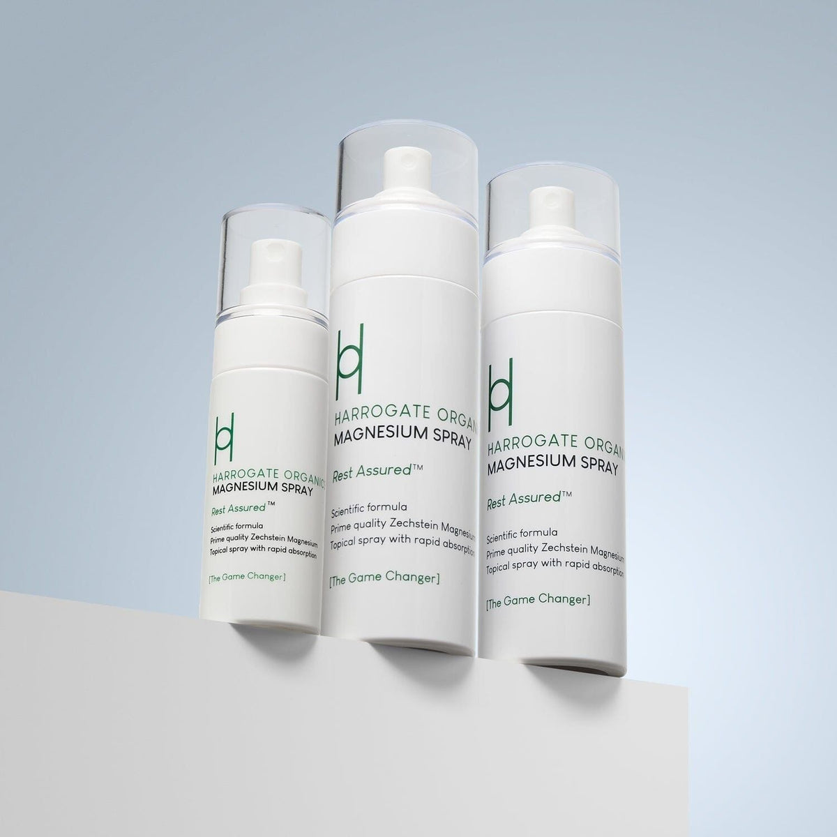 Three white bottles of Harrogate Organics Magnesium Spray - Travel Pack, featuring green text and clear caps, are displayed on a white surface against a light blue gradient background and arranged from smallest to largest.