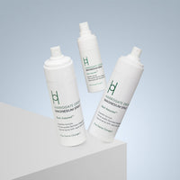 Three white bottles of Magnesium Spray - Travel Pack are shown, two standing upright and one floating, set against a light gray gradient background with a white geometric surface in front.