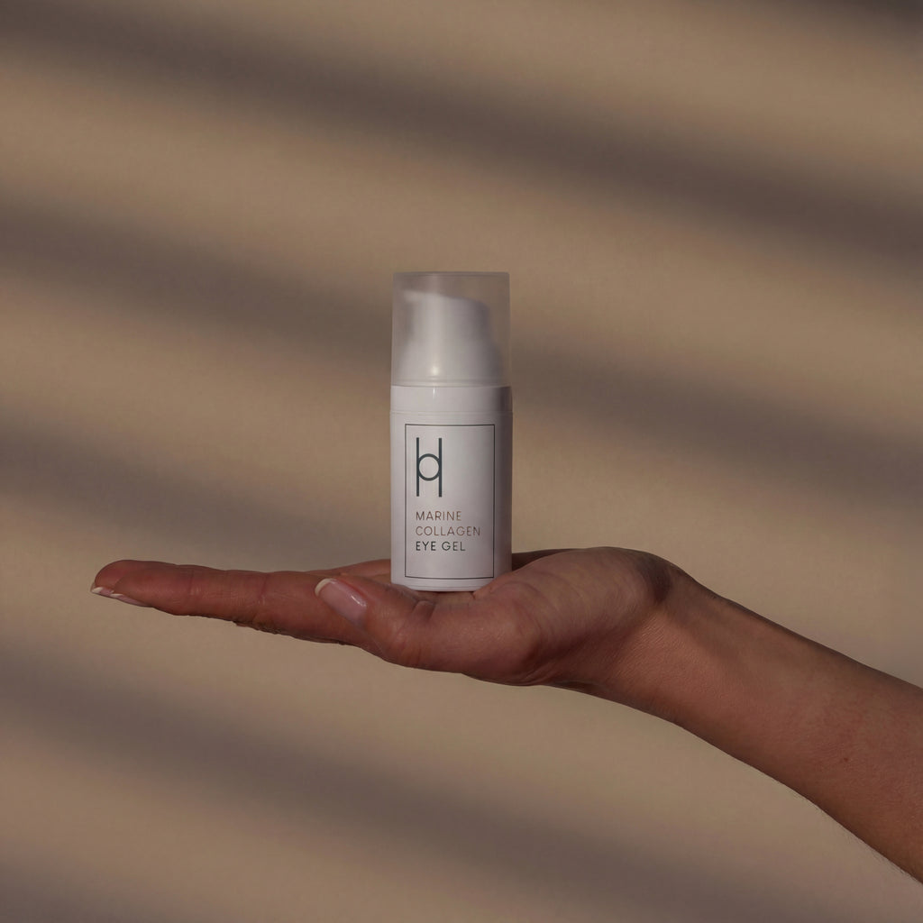 A hand with palm up holds a 30ml white Marine Collagen Eye Gel—formulated with hyaluronic acid, avocado oil & green tea extract to target dark circles—against a beige background with diagonal shadows.