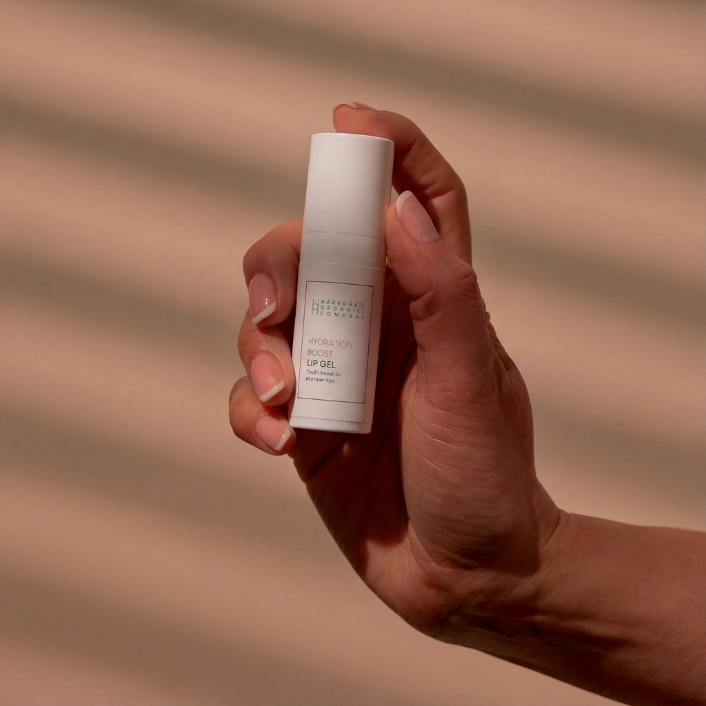 A hand holds a small white bottle labeled “Hydration Boost Lip Gel | Fuller, Hydrated Lips | 10ml” against a background with soft, diagonal brown shadows. The nails are natural and neatly trimmed.