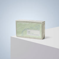 Avo Glow - Luxe Avocado Soap, in pale green floral packaging, rests on a white surface with a light blue backdrop. The label promises clean, nourished skin—enjoy this moisturizing avocado soap as part of your daily routine.