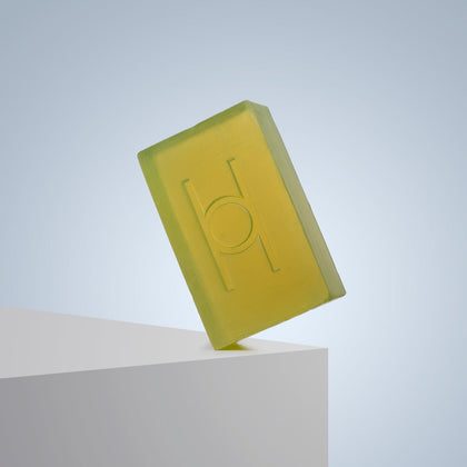 The Avo Glow - Luxe Avocado Soap, a translucent yellow-green bar with an embossed geometric design, is perched on a white edge against a soft blue gradient background.