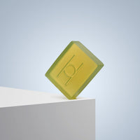 Avo Glow - Luxe Avocado Soap is a translucent yellow-green rectangular bar with an embossed circle and line design, standing upright on a white surface against a light blue gradient background.