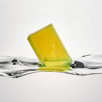 Avo Glow - Luxe Avocado Soap, a translucent yellow-green bar embossed with a "b," floats on clear water against a white background.