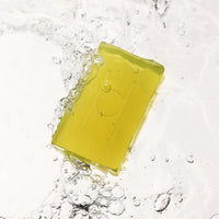 A yellow-green rectangular bar of Avo Glow - Luxe Avocado Soap, featuring an embossed design, is partially submerged in clear water, with splashes and bubbles forming around it.