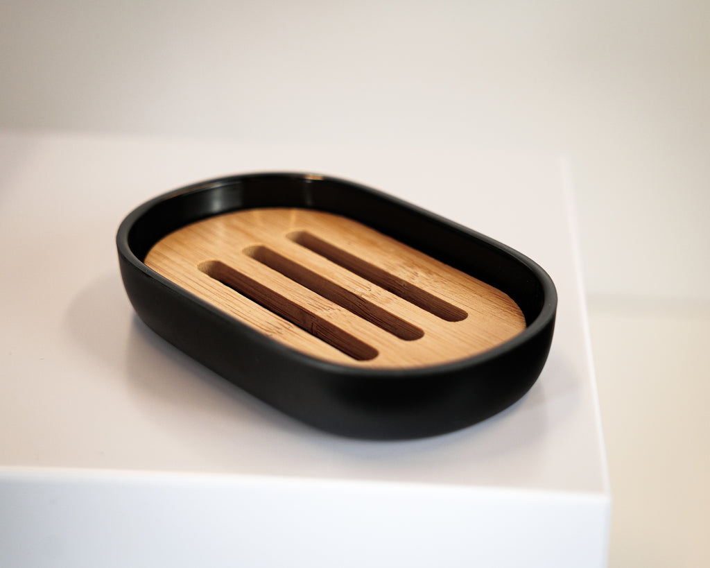 The Sustainable Sugar Cane Soap Dish, featuring black plastic edges and four drainage slots, rests on a white surface.