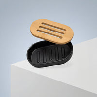 The Sustainable Sugar Cane Soap Dish, oval and black with a matching slatted bamboo lid, rests on a white surface against a light blue background.
