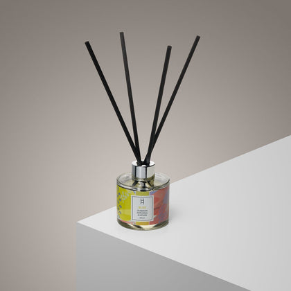 The Bliss Reed Diffuser | Ylang Ylang & Olibanum | 100ml, with black reeds, a silver cap, and a colorful label, sits on a white surface and fills the room with uplifting ylang-ylang and olibanum fragrance.