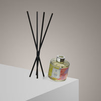 The Bliss Reed Diffuser | Ylang Ylang & Olibanum | 100ml features a clear glass bottle with yellow liquid and black reeds, set on a white surface. Its floral label and natural ylang ylang create an uplifting aroma against a beige gradient background.