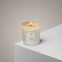 A Bliss Aromatherapy Candle in a glass jar labeled Ylang Ylang & Olibanum burns on a white surface, filling the air with its hand-poured, natural scent against a neutral light beige background.