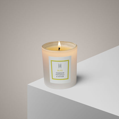 A Bliss Aromatherapy Candle in a glass jar labeled Ylang Ylang & Olibanum burns on a white surface, filling the air with its hand-poured, natural scent against a neutral light beige background.