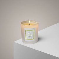A lit white candle in a glass container labeled “Bliss Olibanum Patchouli & Vetiver” sits on the edge of a white surface against a neutral background.