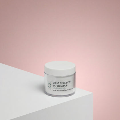 A white jar labeled Stem Cell Body Exfoliator 100ml rests on a white surface with a soft pink gradient backdrop, its minimal modern packaging suggesting the vegan formula inside that gently enhances skin elasticity.