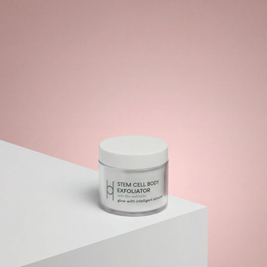 A white jar labeled Stem Cell Body Exfoliator 100ml rests on a white surface with a soft pink gradient backdrop, its minimal modern packaging suggesting the vegan formula inside that gently enhances skin elasticity.