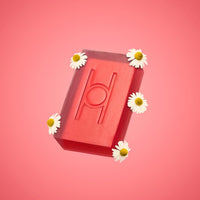 The Calm Chamomile - Luxe Chamomile Soap, a red rectangular bar with an embossed design, is displayed with five white daisies against a pink gradient background.