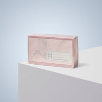 A pink rectangular Luxe Chamomile Soap bar with subtle fish and leaf designs on the packaging rests on a white geometric surface against a light blue background. The label reads, “Calm Chamomile for clean and nourished skin.”.