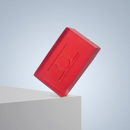 A translucent red rectangular bar, the Calm Chamomile - Luxe Chamomile Soap, featuring an engraved circular and vertical line design, balances on a white surface against a light blue background.