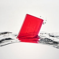 A translucent red bar of Calm Chamomile - Luxe Chamomile Soap with an embossed design splashes into clear water, sending ripples and droplets across a light background.