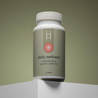 A bottle of Daily Wellness: Multivitamins for Women Under 50 rests on a white platform with a gradient olive background. The green label features energy and immune support, plus a pink, flower-like icon in a minimalist design.