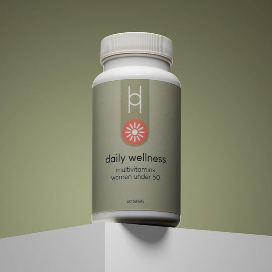 A bottle of Daily Wellness: Multivitamins for Women Under 50 rests on a white platform with a gradient olive background. The green label features energy and immune support, plus a pink, flower-like icon in a minimalist design.