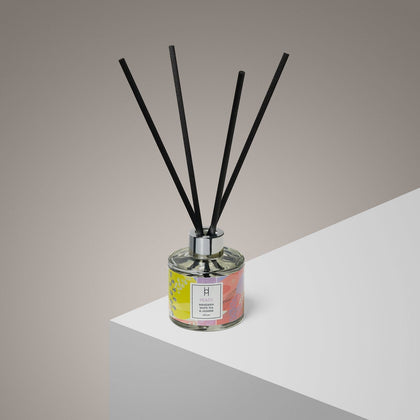 The Peace Reed Diffuser | Mandarin & Jasmine | 100ml, with four black reeds, a silver cap, and a colorful label, rests on a white surface against a neutral background, filling the room with its natural fragrance.