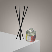 The Peace Reed Diffuser | Mandarin & Jasmine | 100ml features a glass bottle with a colorful label on a white surface, paired with five black diffuser sticks. The beige-to-gray background evokes calm, enhancing your space with natural fragrance.