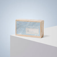 A rectangular bar of Elixir Edition Luxe Soap wrapped in light blue and beige paper with an abstract leaf pattern, displayed on a white pedestal against a pale blue background. Label: Elixir Edition for intense moisturisation and nourished skin.