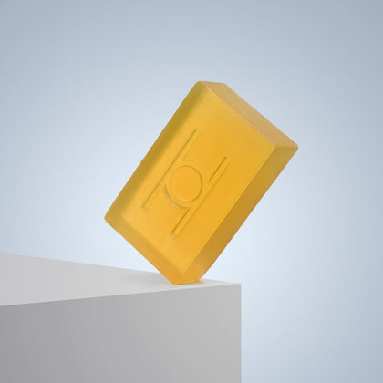The Surprise & Delight Free Soap Bar, a translucent yellow vegan soap with a geometric design, is balanced on the edge of a white surface against a light blue gradient background.