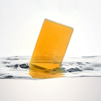 A translucent amber Surprise & Delight Free Soap Bar, partially submerged at an angle in clear water, displays a simple embossed logo on its surface against a white background.