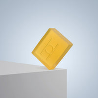 A translucent yellow Elixir Edition Luxe Soap bar with an embossed circular design balances on the edge of a white surface against a light blue gradient background.
