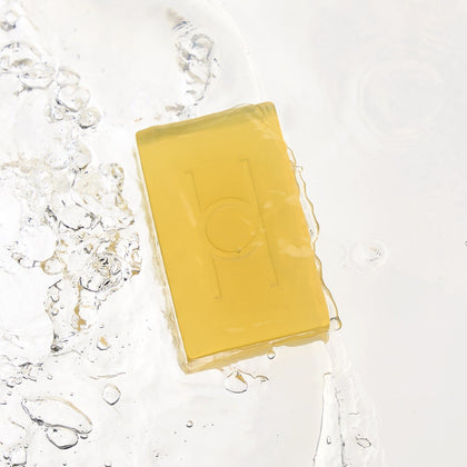The Surprise & Delight Free Soap Bar, featuring a translucent yellow hue and simple geometric design, is shown partially submerged in clear water, surrounded by bubbles and gentle ripples.