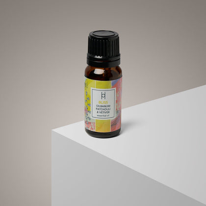 A small brown glass bottle with a black cap labeled Bliss Essential Oil Blend | Ylang Ylang & Olibanum | 10ml stands on a white and gray geometric surface against a neutral background.