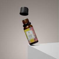 A small amber glass bottle labeled “Bliss Essential Oil Blend | Ylang Ylang & Olibanum | 10ml” with a colorful label floats above a white platform, its black cap hovering above, highlighting this aromatherapy oil against a neutral background.